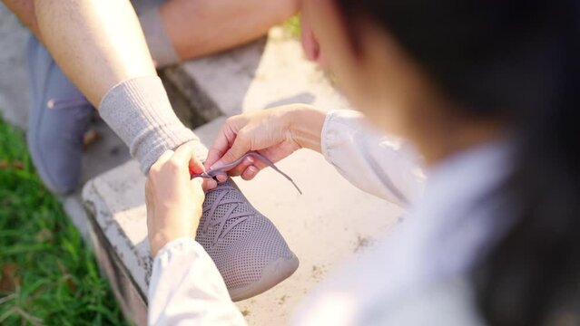 Asian woman daughter helping elderly father tie running shoe laces during jogging exercise together at park. Retired senior man health care with outdoor lifestyle sport training workout in the city.