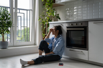 Unhappy Asian girl sitting on floor in kitchen feeling depressed after social media usage, being ignored by boyfriend, dealing with rejection in love. Breakup depression concept - obrazy, fototapety, plakaty