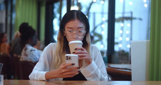 Asian woman in eyeglasses sitting at cafe and scrolling smartphone app using mobile internet. Asian female student with smartphone, drinking hot coffee at modern cafe. People using smartphone concept