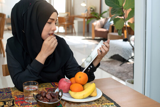 Image of Muslim Woman wearing black hijab holding digital tablet  while eating dry date fruit at home, plate of fruit was placed on the table in front of her