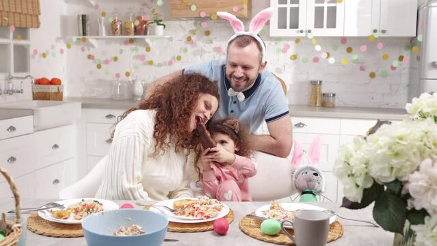 Portrait of loving family celebrating holy holiday in beautifully decorated domestic kitchen. Cute baby girl eating chocolate Easter bunny with her parents. High quality 4k footage