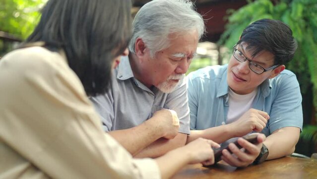 Happy Asian couple teaching elderly father using mobile phone application during having lunch together at restaurant on summer vacation. Family relationship older people mental health care concept.