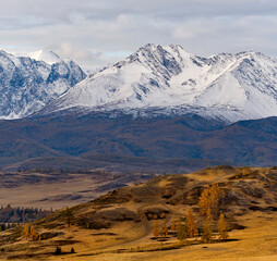 Russia, the Altai Mountains. View of the autumn mountains with a winding dirt road stretching into the distance against the background of the snow-covered North Chui mountain range.