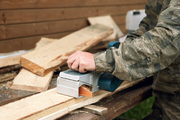 A man in military uniform works with an electric planer tool on the street, grinds wooden boards...