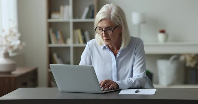Aged businesswoman sits at desk working on laptop in cozy home office room, looks at device screen, learn received e-mail, ponder on task, doing on-line job, prepare report. Business, tech, workflow