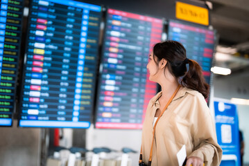 Young asian woman with passport and boarding pass as a hand in international airport looking at the flight information board, checking her flight
