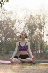 Relaxed woman enjoying music while doing yoga exercises in the public park. The young lady feels free, relaxed, and happy.