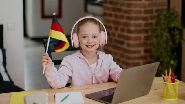 Cute little girl wearing headset studying online on laptop, shaking Germany flag and smiling to camera, free space