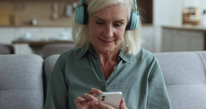 Close up blissful mature retired woman relaxing at home, puts on modern wireless headphones, play favourite music using smartphone. On-line audio digital streaming services user, modern tech, hobby