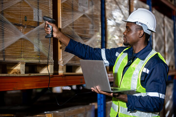 Fototapeta na wymiar Black man professional worker wearing safety uniform and hard hat using laptop and scanning box inspect product on shelves in warehouse. Male worker is pointing finger inspecting product in factory.