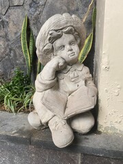 Boy sculpture crafted from cement looks  thinking  imagine holding book with plants and rock wall at the background. An ornamental monument of human creativity and representation.