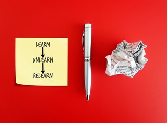 Pen, crumpled paper and note paper on red background with handwritten text Learn Unlearn Relearn -...