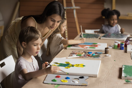 Friendly young art teacher giving artistic lesson to kids, helping to draw colorful cartoon animals, blend colors on palette, painting on canvas. Diverse group of children training creativity