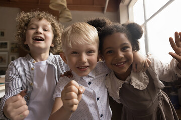 Cheerful diverse kids celebrating studying success, hugging in classroom, smiling, laughing, making winner hands, looking at camera, smiling, laughing, having fun, enjoying school friendship