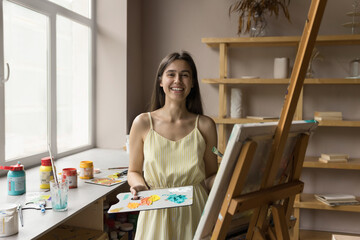 Happy beautiful young art school student drawing on canvas at easel, using artistic supplies,...