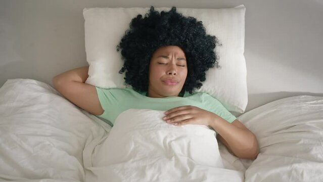 Top down view above female waking up on early morning in comfy bed, yawns and closing open mouth with palm. Afro girl wants to sleep lying on comfortable pillow under blanket in slow motion on morning