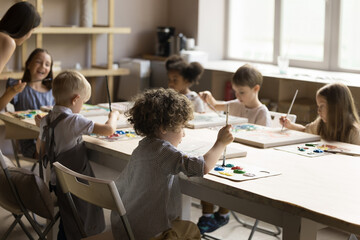 Group of little artistic school pupils drawing in acrylic colors on class, studying art under supervision of teacher, mixing acrylic paints on palettes, sitting at big table with canvas