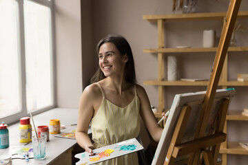 Happy dreamy student girl drawing in acrylic paints in art school, stroking on canvas, looking...