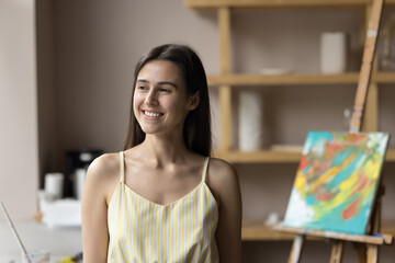 Happy dreamy young artist woman enjoying creative hobby, standing in art studio, looking at window away with inspiration, thinking over idea for artwork, smiling