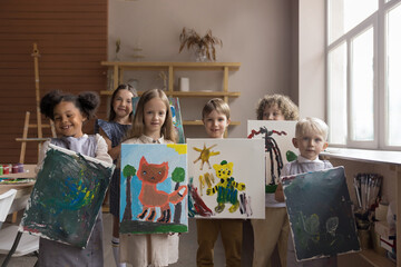 Diverse group of happy proud kids attending artistic school class, standing together, holding canvas, showing painted pictures, looking at camera, smiling, laughing, studying creativity, art