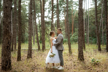 young couple bride in a white short dress and groom in a gray suit in a pine forest