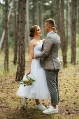 Fototapeta premium young couple bride in a white short dress and groom in a gray suit in a pine forest