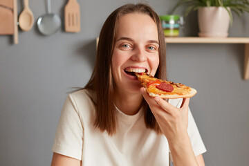 Portrait of smiling adorable dark haired woman wearing white t shirt posing in kitchen and eating...