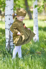 Portrait of a cute little girl in a uniform among birches on a green background. Victory Day, May 9 holiday.