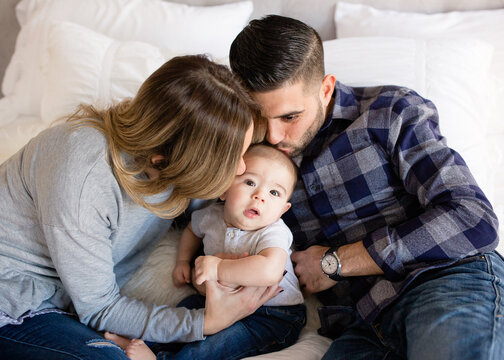 Mom and dad kissing cute baby boy on cheek indoors on bed