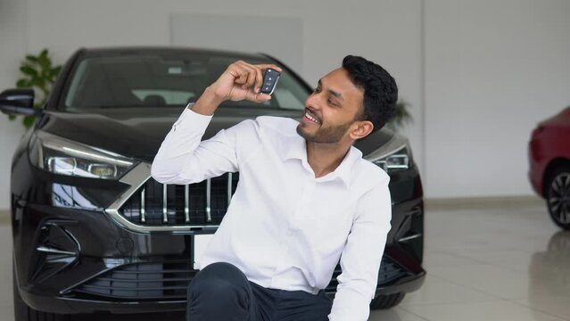 Positive pakistani male smiling for camera and demonstrating keys while sitting near new vehicle in showroom