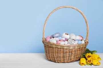 Wicker basket with festively decorated Easter eggs and beautiful tulips on white wooden table against light blue background. Space for text