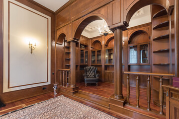 A luxurious office in a private house, decorated with natural dark wood. Bookcases, shelves and a comfortable armchair. - obrazy, fototapety, plakaty