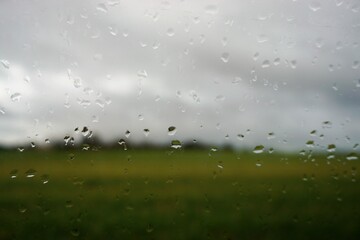 Abstraktes Motiv mit Glasscheibe und Regentropfenmuster vor gr&uuml;nem Feld, B&auml;umen und grau-wei&szlig;em Himmel bei Regen und Sturm am Morgen im Winter
