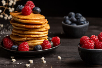 Close-up of plate with stacked pancakes, blueberries and raspberries, on table and dark background with white flowers, selective focus, horizontal. with copy space