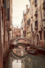 Arched bridge over a narrow canal in Venice with old houses and parked boats next to them (vintage style photo)