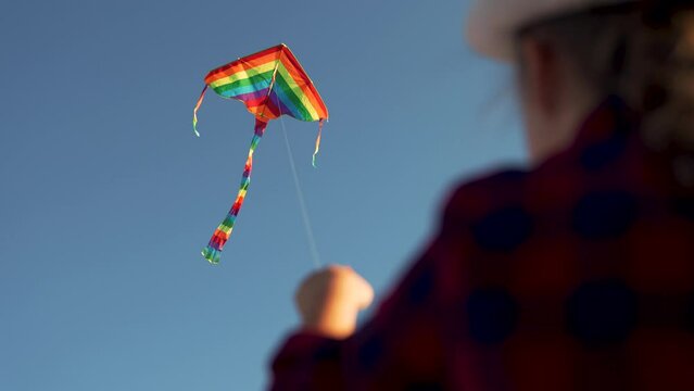 Kite soars in wind in sky. Child is holding toy kite. Girl in grass in park. Colored toy in sky. Child hand with toy kite. Girl is playing in park. Child hand with kite soaring in wind.