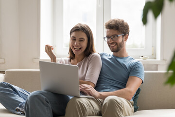 Joyful millennial couple in love talking on video call on laptop, enjoying online, internet communication, leisure at computer, relaxing on home sofa, hugging, watching movie, laughing