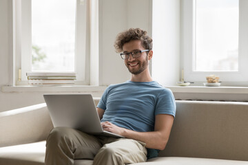 Positive successful handsome freelance business man sitting on sofa, using laptop, typing, chatting, smiling, laughing, looking at camera, posing for portrait. Millennial employee man working at home