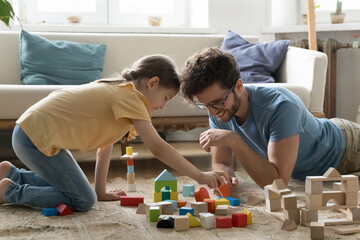 Positive engaged young dad and daughter kid playing with cubes on carpeted floor, building toy...