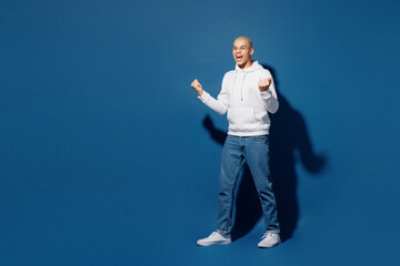 Full body side view young man of African American ethnicity wear white hoody doing winner gesture celebrate clenching fists say yes isolated on plain dark royal navy blue background studio portrait.