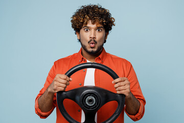 Young shocked amazed astonished Indian man wear orange red shirt white t-shirt hold steering wheel driving car isolated on plain pastel light blue cyan background studio portrait. Lifestyle concept.