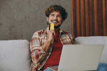 Young Indian man wear casual clothes use mobile cell phone hold credit bank card shopping online sits on grey sofa couch stay at home hotel flat rest relax spend free spare time in living room indoor.