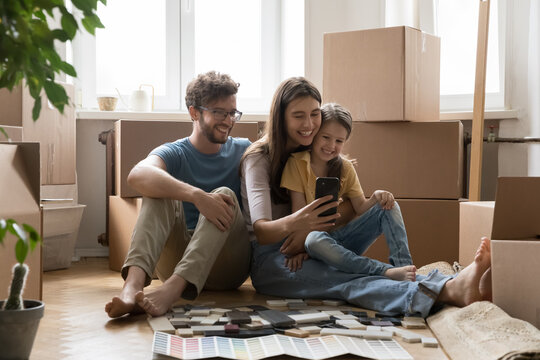 Positive Parents And Happy Kids Taking Moving Selfie On Smartphone, Sitting At Stacked Cardboard Boxes, Renovation Brochures With Wall Color Samples, Holding Mobile Phone, Smiling