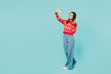 Full body young woman wearing bright casual clothes doing selfie shot on mobile cell phone post photo on social network show v-sign isolated on plain pastel light blue cyan background studio portrait.