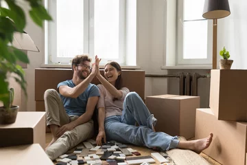 Fotobehang Cheerful excited young couple planning renovation after moving into new apartment, choosing interior material samples of tile, laughing, giving high five, clapping hands at cardboard boxes © fizkes