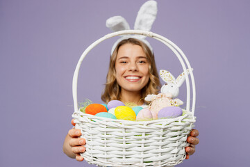 Close up young cheerful fun woman wearing bunny rabbit ears hold wicker basket colorful eggs look aside on area isolated on plain pastel light purple background studio portrait. Happy Easter concept.