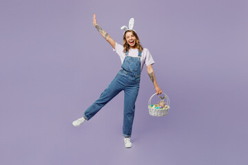 Full body smiling fun young woman wearing casual clothes bunny rabbit ears holding wicker basket colorful eggs raising hand up isolated on plain pastel purple background studio. Happy Easter concept. - obrazy, fototapety, plakaty