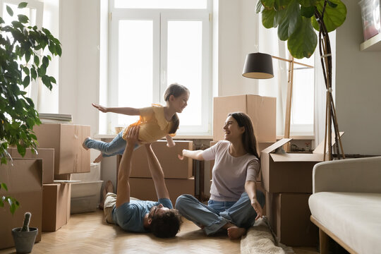 Cheerful positive parents and little child celebrating moving into new home, playing on floor at heap of paper boxes. Dad lifting girl with open arms up in air, playing airplane flight
