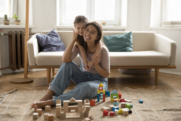 Joyful sweet child embracing happy pretty young mom sitting on floor at toy castle. Positive mother and cheerful kid looking at camera, posing, playing game wit building blocks at home