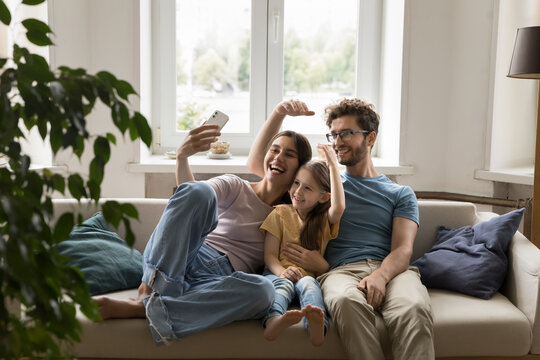 Cheerful happy family taking selfie on couch at home, resting on sofa with smartphone, hugging, smiling, laughing, making hand house gesture, imitating roof, rooftop overhead